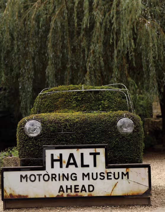 car covered in moss with a sign in front that reads "Halt, motoring museum ahead"