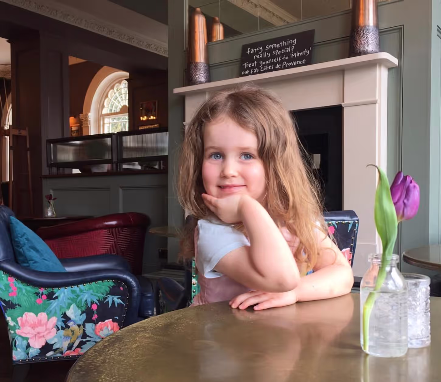 little girl sitting at a wooden table smiling and posing with her head in her hand