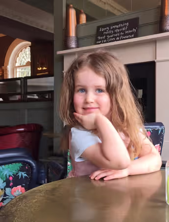 little girl sitting at a wooden table smiling and posing with her head in her hand