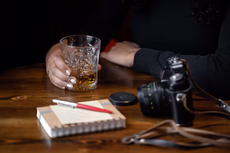 person holding a glass of liquor on a table next to a camera and a notepad