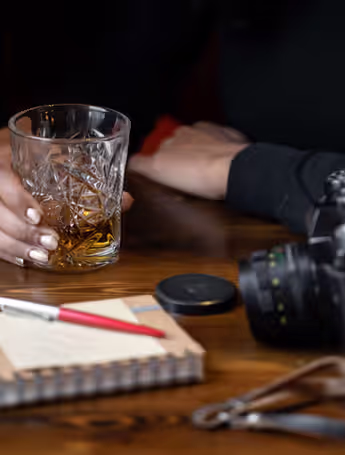 person holding a glass of liquor on a table next to a camera and a notepad
