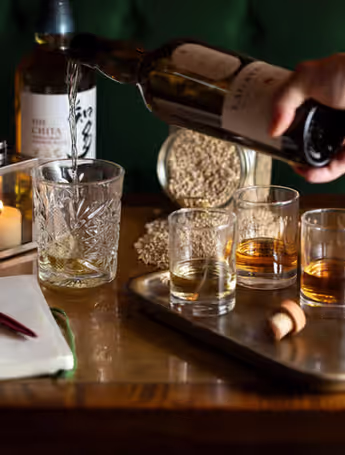person pouring whiskey into a glass next to a tray of three whiskey glasses and a notepad